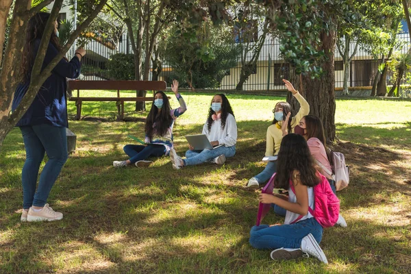 Masked Latina teenage students raise their hands during an outdoor class with a teacher in a park