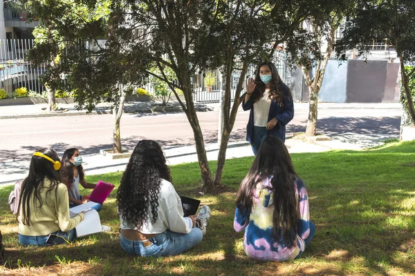 Latina teacher teaching in a face mask with her teenage students with their backs to the lesson in an outdoor classroom.