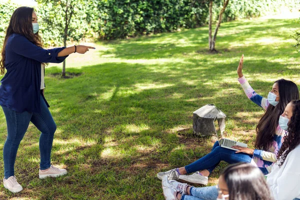 Latina teacher gives the floor to a student during an outdoor class, they wear a mask and social distancing as prevention against the coronavirus during the pandemic