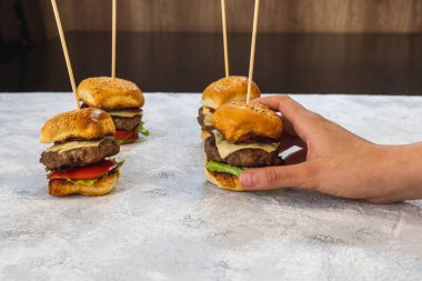 Hafif bir yüzeyde mini bir çizburger, pastırma, marul ve domates alan bir elin yatay görüntüsü.