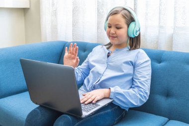 Sarışın kız mavi bir koltukta dizüstü bilgisayarıyla video konferansına el sallıyor.