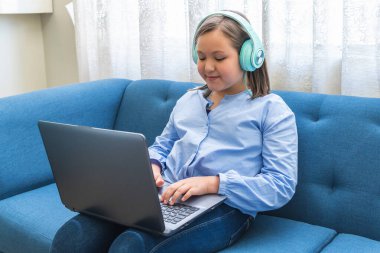 Oturma odasındaki kanepede dizüstü bilgisayar kullanarak gülümseyen kulaklı sarışın kız.