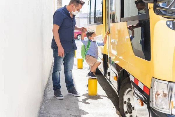 Child with mask getting on the school bus accompanied by his father