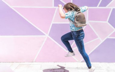 Okula giderken sırt çantalı sarışın bir kız pembe ve mor bir duvarın önüne atlıyor.