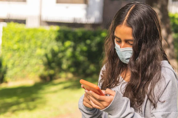 Maskeli Latin esmer kız parkın içinde cep telefonunu kullanıyor.