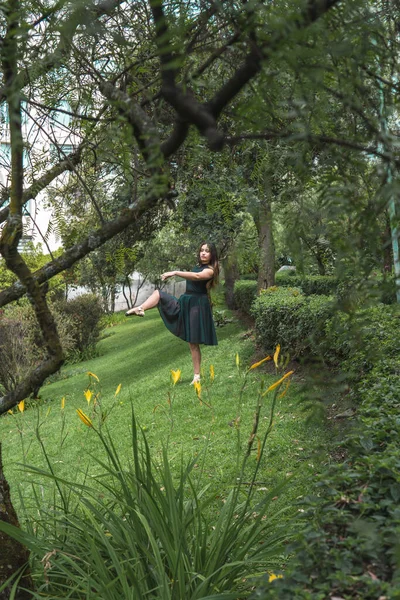 Latin teenage girl in black dress and pointe shoes practicing ballet in a park amidst the greenery