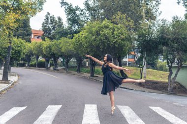 Siyah elbiseli genç balerin kız bale yapıyor. Şehrin caddesinde bir zebranın kesiştiği yerde bale pozu veriyor.