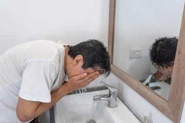 Beyaz, modern bir banyoda yüzünü temiz suyla yıkayan bir adam.