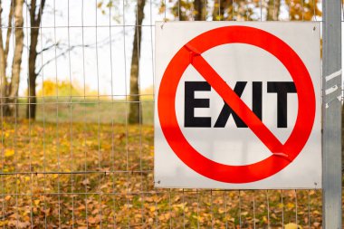 Metal bir tel örgüde yasaklama işareti. Kırmızı, beyaz arkaplanda EXIT yazısıyla daire çizmiş. Arka planda sarı yapraklı bir park var. Sonbahar günü. Boşluğu kopyala.