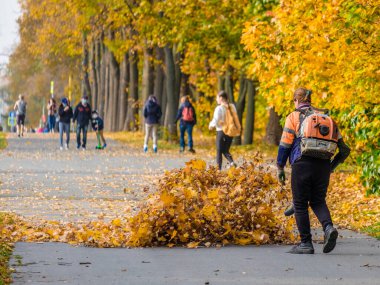 Moskova. - Rusya. 11 Ekim 2020: Bir kadın elektrik işçisi parktaki bir ara sokakta düşen yaprakları temizlemek için hava üfleyici kullanıyor. Sarı yapraklar havada uçuşuyor. Sonbahar güneşli bir gün. Mevsimlik iş kavramı.