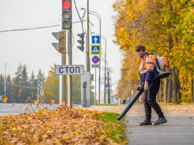 Moskova. - Rusya. 11 Ekim 2020: Bir kadın elektrik işçisi parktaki bir ara sokakta düşen yaprakları temizlemek için hava üfleyici kullanıyor. Sarı yapraklar havada uçuşuyor. Sonbahar güneşli bir gün. Mevsimlik iş kavramı.
