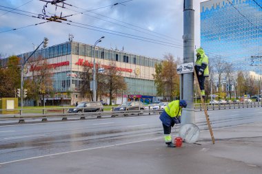 Moskova. - Rusya. 19 Ekim 2020 Sarı üniformalı iki işçi bir sokak lambasının üzerinde yol işareti oluşturuyorlar. Bir adam merdivenin üzerinde duruyor. Bulutlu bir sonbahar günü. Arabalar yol boyunca gidiyor..