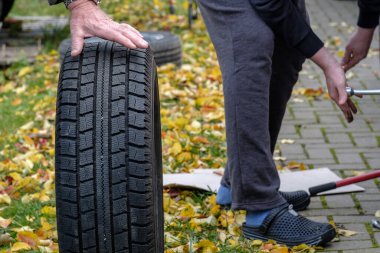The man's hands are holding a new black rubber tire. Seasonal tire change. Autumn day. Yellow fallen leaves on the ground in the background. clipart