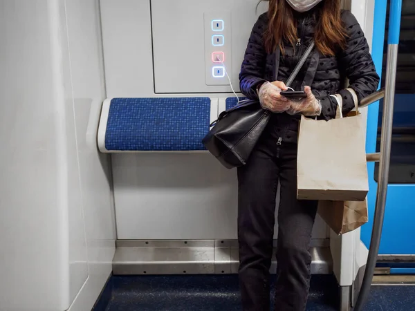 地下鉄の車の中で若い女性がスマホを持っている 乗客の顔には保護マスクが付いている 女性はセロハン手袋をしている 公共交通機関におけるウイルス感染からの保護 — ストック写真