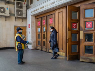 Yüz maskesi takan ve üniforma giyen bir adam metro istasyonundan çıkan insanlara gazete dağıtıyor..