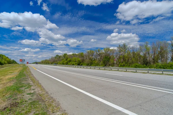 A wide asphalt highway with road markings against a blue sky with clouds.