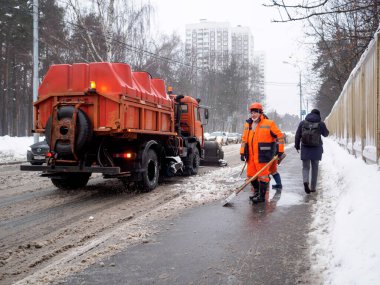 Moskova. - Rusya. 25 Şubat 2021. Turuncu üniformalı kamu görevlileri yoğun bir kar yağışı sonrası çamurlu karı kaldırımdan kürekle temizliyorlar. Sıkı çalışma. Bulutlu kış günü.