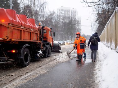 Moskova. - Rusya. 25 Şubat 2021. Turuncu üniformalı kamu görevlileri yoğun bir kar yağışı sonrası çamurlu karı kaldırımdan kürekle temizliyorlar. Sıkı çalışma. Bulutlu kış günü.