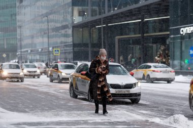 Moscow. Russia. February 12, 2021. A woman in a fur coat and a medical mask with a smartphone in her hand walks in winter along a city street against the background of many taxi cars. Snowing. 