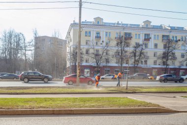 Turuncu üniformalı hademeler şehir caddesinde yol boyunca çimleri süpürüyor..