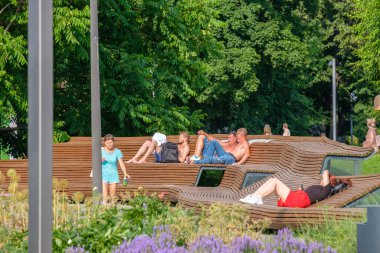 Yarı çıplak insanlar güneşlenir ve rahatlarlar. Parktaki ahşap banklarda uzanırlar..