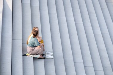 İki genç kadın, büyük bir merdivenin gri taş basamaklarında yalınayak oturuyor..