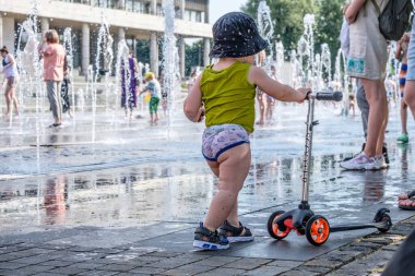 Şapkalı küçük bir çocuk fıskiyelerin arka planında scooter tutuyor.