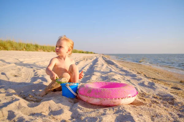 Seorang bayi perempuan pirang kecil bermain di pasir di pantai pada hari yang cerah. — Stok Foto