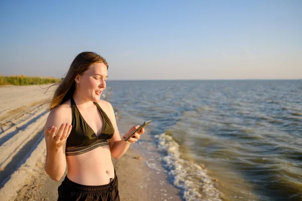 Deniz kıyısındaki genç bir kadın akıllı telefon kullanarak iletişim kuruyor.. — Stok fotoğraf
