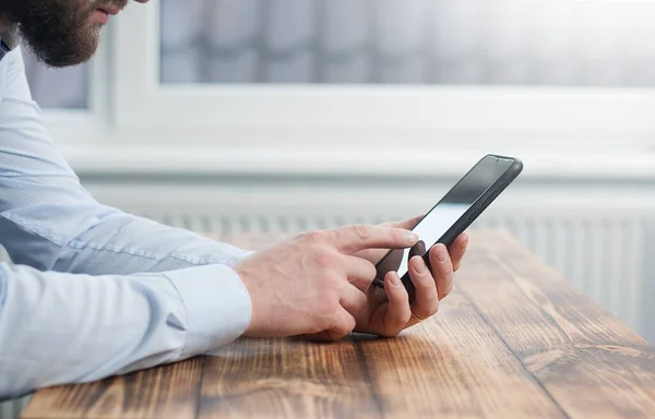 Young male businessman holding a phone in his hands touching the screen. Sitting at a wooden table