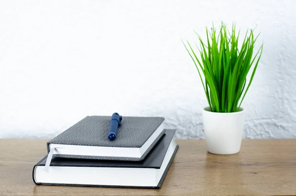 Closed book and notepad and pen. Green flowerpot in a white pot on a wooden table. Desktop.