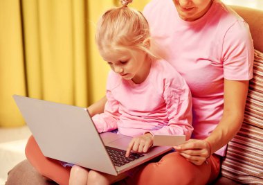 Çevrimiçi alışveriş veya ödeme konsepti. Annem 4-5 yaşındaki kızına evde bilgisayar ve kredi kartı kullanıyor. Pembe elbiselerle kanepede oturuyor.