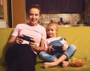 Happy family together. Mother and her child girl playing video games sit on couch ahd hold gamepad. People having fun at home clipart