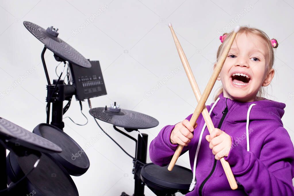 Happy caucasian kid girl drummer. Girl playing on elettronic drum kit or learns to play drums in music school. Emotional portrait. Mock up, studio on gray background. Free space for advertisement.