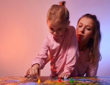 Aile bir masa oyununda evde birlikte oynayarak eğleniyor. Oyun alanı, bir sürü figür, bozuk para ve kum saati. Anne ve tatlı küçük kız oynuyorlar, ellerinde kartlarla neşeli duygularla..