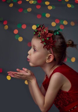 Noel öpücüğü. Kırmızı pullu elbise ve Noel çelengi giyen kız çocuğu. Sarı saç ve mavi gözler stüdyoda gri arka planda ışık ve sahte ışıklarla poz veriyor ve maskaralık yapıyor..