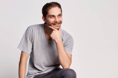 Tişörtlü yakışıklı genç adam sırıtarak sandalyede oturuyor ve elini yüzüne yakın tutuyor. Gri arka planda izole bir şekilde.