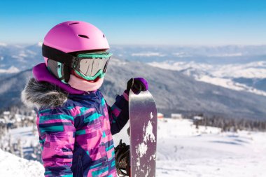 Kar kayağı yapan küçük bir kız, gözlüklü ve teçhizatlı güneş ışığı altında kar kayağı yapıyor.