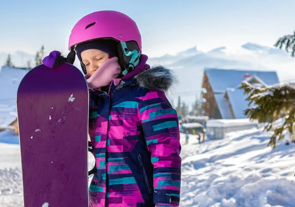 Little cute girl snowboarding at ski resort in sunny winter day. Caucasus Mountains. Portrait of kid in helmet and snowboard in winter resort