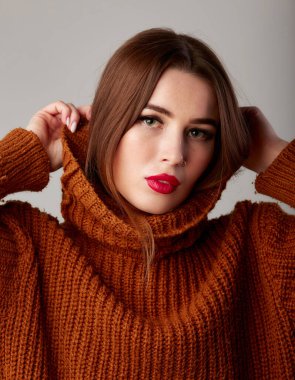 Balıkçı yaka kazaklı, yaka kazaklı genç ve güzel bir kadın. Kırmızı dudaklı sevimli bir kadın model. Stüdyoda, gri arka planda.