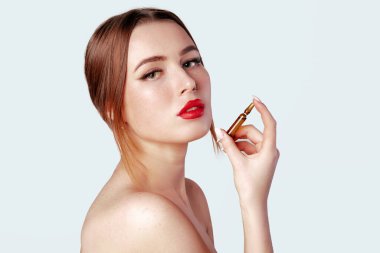 Kadının elinde saç veya cilt bakımı için serum var. Burun küpeli, kırmızı dudaklı ve gri arka planda mükemmel makyajlı çekici bir kadın fotoğrafı. Güzellik konsepti