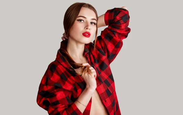 Beautiful face of woman with nose earring in red plaid shirt. Portrait of cute brunette with red lips and nose piercing posing in a red checkered shirt on gray