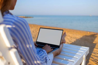 Boş masaüstü ekranı olan bir dizüstü bilgisayar kullanan ve deniz kenarında mavi gökyüzü arka planında oturan bir kadının görüntüsü. Serbest çalışma konsepti. Teknoloji ve seyahat.