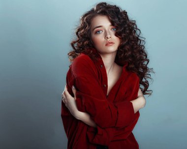 Uzun kıvırcık saçlı çekici bir esmer. Kırmızı gömlekli, dalgalı saçlı, mavi arka planda poz veren kadın model.