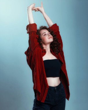 Uzun kıvırcık saçlı çekici bir esmer. Kırmızı gömlekli, dalgalı saçlı, mavi arka planda poz veren kadın model.