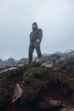 Yaz yağmurunun altında, dağın tepesinde su geçirmez ceketli yakışıklı, sakallı bir adam. Doğal arkaplanda bulutlu gökyüzüne karşı dağ tepesi, kopyalama alanı