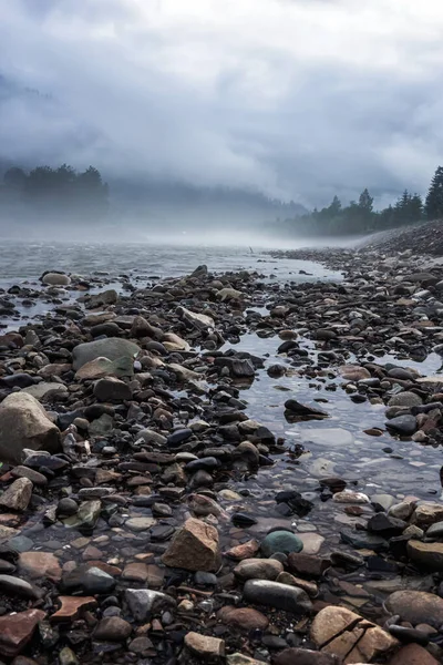 Dark and moody river and rocks landscape with fog in the trees and water. Moody nature vibes with mist and dark cold weather.