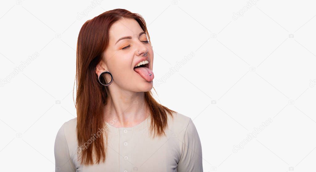 Emotions woman showing tongue. Having earrings in the nose sticking out her tongue and showing piercing. White background, copy space. Woman face piercing