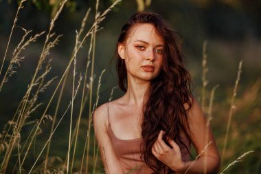 Beautiful woman face stand against wild grass, forest and bush in sunny rays field at sunset in summer meadow. Alone Cute young adult woman enjoy golden hou warm evening