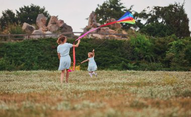 Young mom with a little daughter play with flying a kite in a green park happy family having fun together. Free space for text